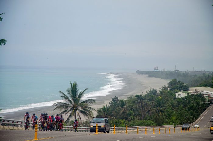 Nádherné pobřeží v pozadí při právě probíhajícím odlivu. Na silnici u pobřeží skupina cyklistů právě bojuje s převýšením.