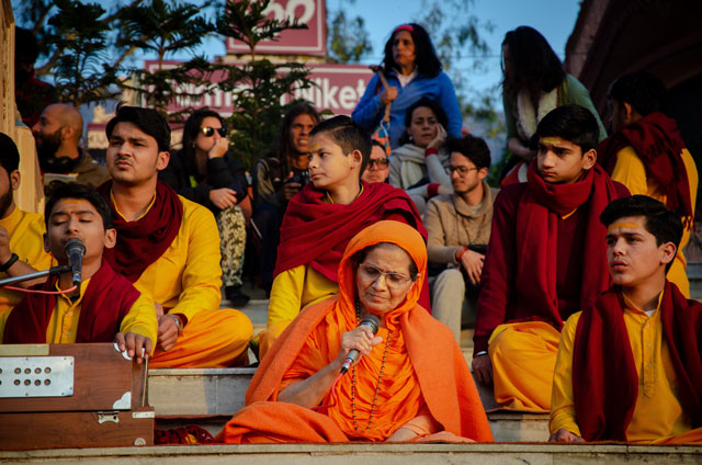 Uprostřed sedí stará žena, která vystupuje jako učitelka jógy a právě zpívá mantry během obřadu aarti. 
