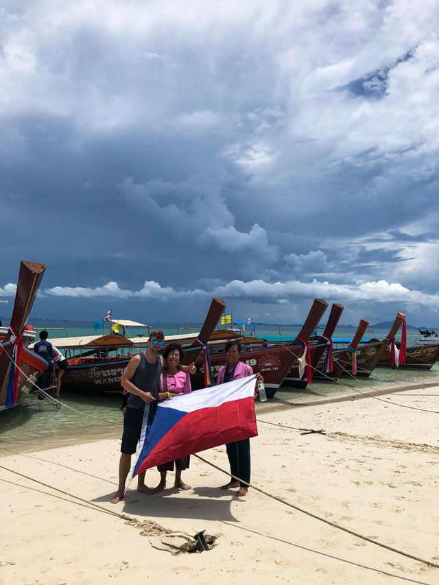 Já, společně s dvěma tajkami, držící českou vlajku. Jsme na jednom z ostrovů Krabi a za námi, jsou známe Thajské loďky.